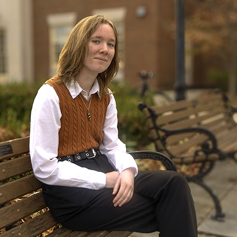 Marlow, a student wearing a tan sweater vest, white shirt, and black pants, sits on one of the wooden benches outside King Library.
